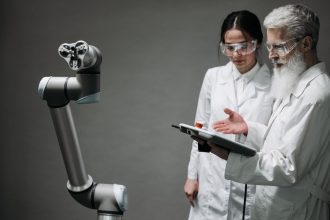 Pesquisadores usando jalecos e óculos de proteção analisam um braço robótico em um laboratório.