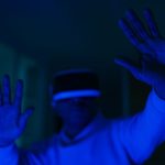 Homem usando headset de realidade virtual e interagindo com as mãos em um ambiente iluminado por luz azul.